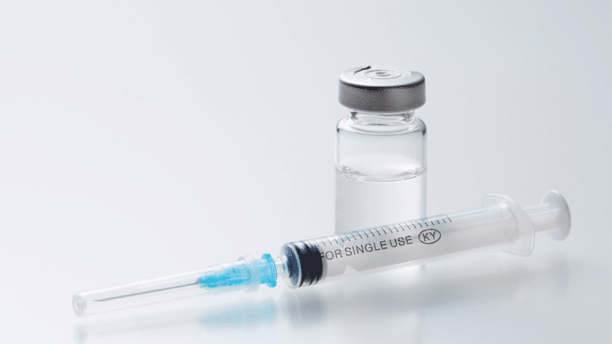 A close-up shot of a clear medical syringe with a blue needle hub and cap, lying on a reflective white surface next to a glass vial filled with clear liquid and topped with a silver cap.