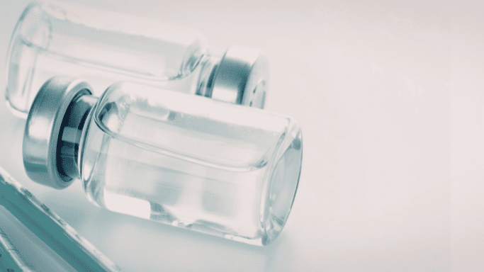 A high-angle, close-up shot of two glass medication vials with silver caps lying on a white surface, with the tip of a syringe visible in the lower-left corner. The image has a cool, blue-toned filter.