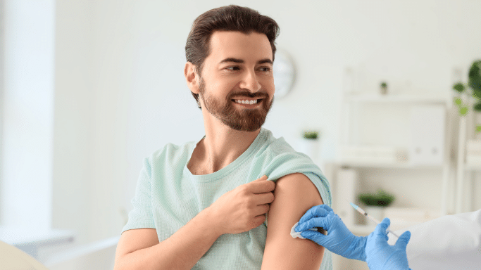 A smiling man in a light green t-shirt sitting in a bright clinic setting while a healthcare professional, wearing blue nitrile gloves, prepares his upper arm for an injection with a cotton swab.