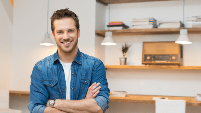 A smiling young man with short brown hair and a light beard stands with his arms crossed in a modern, brightly lit room. He is wearing a blue denim button-down shirt over a white t-shirt and a silver watch.