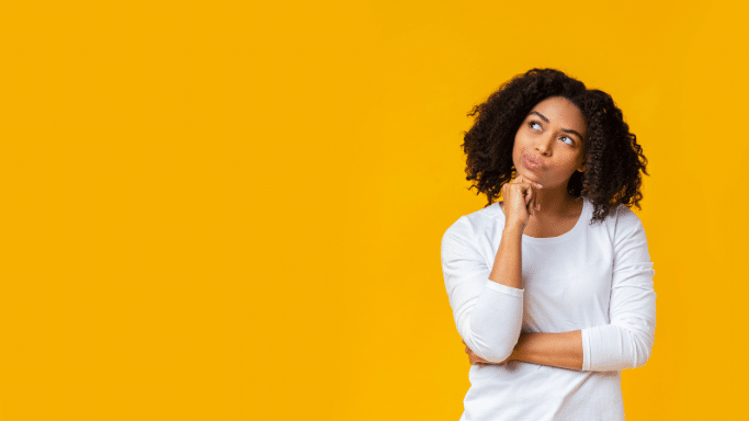 woman with curly black hair stands against a solid bright yellow background. She has a thoughtful expression, with her hand on her chin and her eyes looking upward and to the side as if she is contemplating an idea.