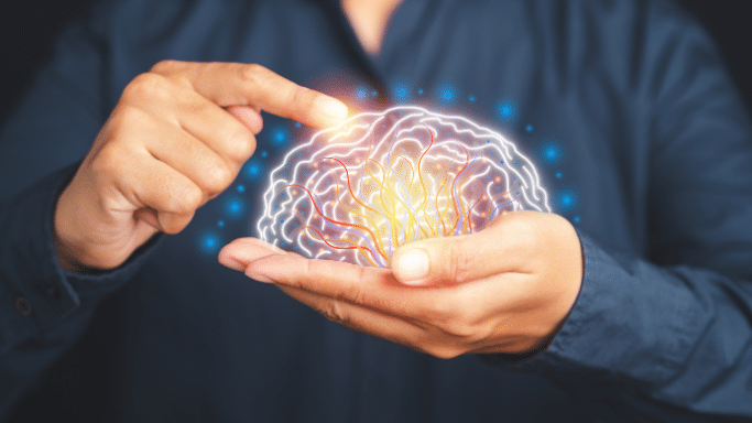 A person’s hands cupping a glowing, stylized white outline of a human brain. Inside the brain, vibrant orange, yellow, and blue neural lines pulse with light. One finger points toward a bright spark at the top of the brain against a dark, out-of-focus background.