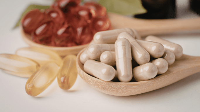 A close-up of two light-colored wooden spoons resting on a white surface. One spoon is filled with tan-colored capsules, while the spoon in the background holds reddish-brown translucent softgels. A few yellow softgels are scattered on the surface to the left.