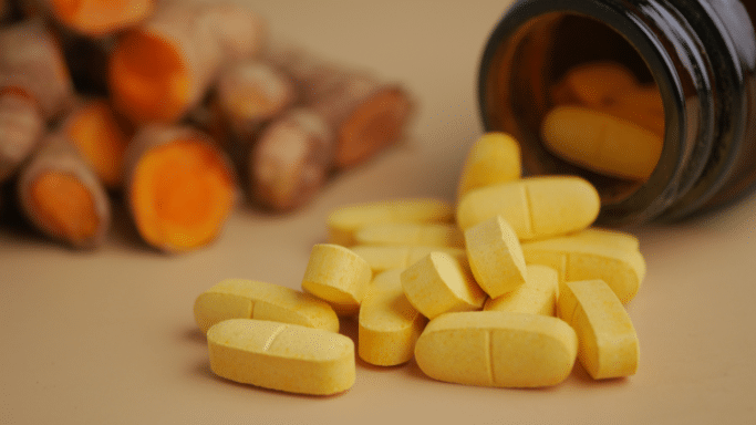 A pile of bright yellow oval tablets spilling out of a dark amber glass bottle. In the soft-focus background, several pieces of raw turmeric root are sliced open, revealing their deep orange interior, emphasizing a natural or herbal connection.