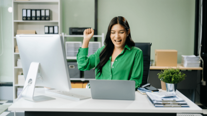woman with long dark hair sits at a white office desk, cheering with a raised fist and a joyful expression. She is wearing a vibrant green button-down shirt. On her desk are a large desktop monitor, a laptop, and various office supplies, set against a background of white bookshelves and a light green wall.