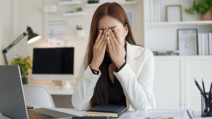 A woman in a white blazer sits at a desk, pressing her fingers to the bridge of her nose with her eyes closed in an expression of stress or fatigue.
