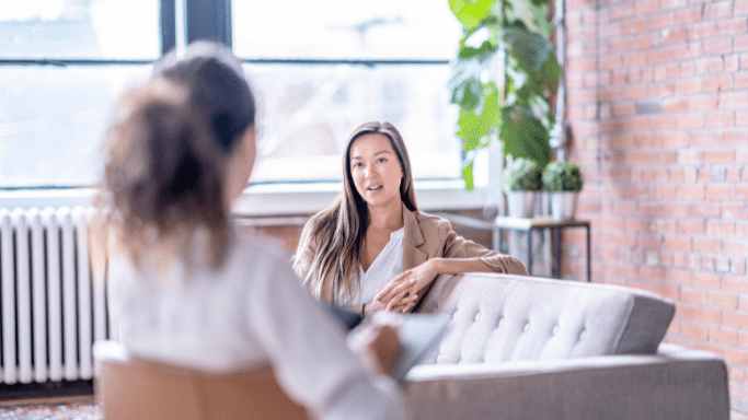 A woman in a tan blazer sits on a grey sofa, engaged in a conversation with another person whose back is to the camera. The setting is a bright, modern office or studio with large windows and a red brick wall in the background.
