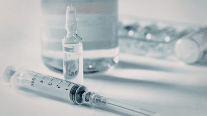 A detailed close-up of medical equipment, featuring a clear glass ampoule standing upright next to a plastic syringe. In the blurred background, several more vials are visible. The image has a clean, clinical blue-grey tint.