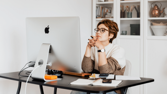 A woman with short brown hair and glasses sits at a black desk, looking thoughtfully at a large silver desktop monitor. She has her hands clasped under her chin in a pensive pose. The room is bright with white shelving in the background.
