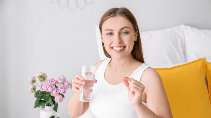 A smiling woman sitting in bed holding a glass of water and a small white pill.
