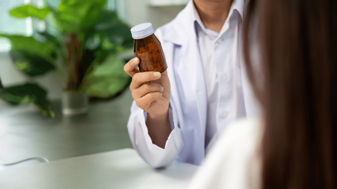 A doctor in a white coat holding up an amber medicine bottle while speaking to a patient.