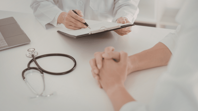 An overhead view of a medical consultation featuring a stethoscope, a laptop, and a doctor writing on a clipboard while a patient's hands are clasped.