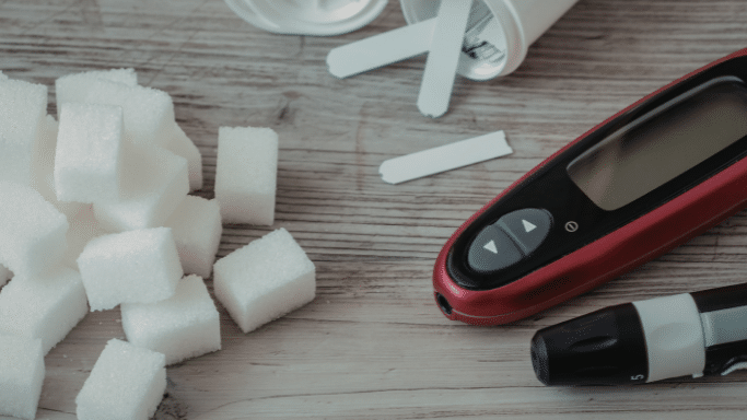 A pile of white sugar cubes sits next to a digital glucose monitor, test strips, and a lancing device on a wooden surface.