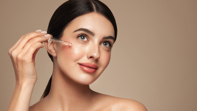 A close-up of a woman with dark hair and blue eyes applying a clear liquid from a glass dropper onto her cheek. She has glowing, clear skin and is looking upward with a slight smile against a neutral beige background.