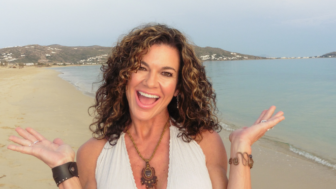 A joyful woman with curly brown hair and bohemian jewelry smiles broadly on a beach with her arms outstretched in excitement.
