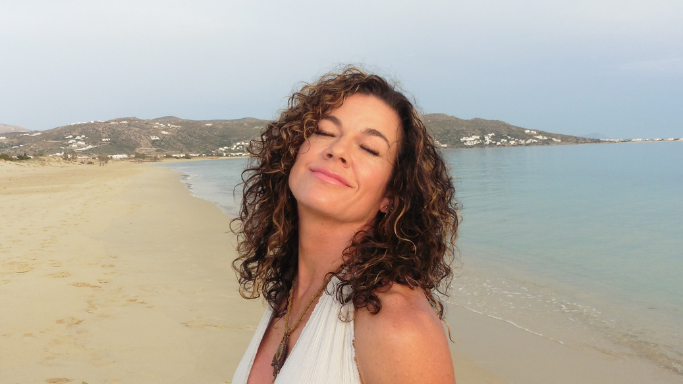 A peaceful close-up of the woman on a beach, eyes closed and head tilted back with a gentle smile against a soft coastal background.