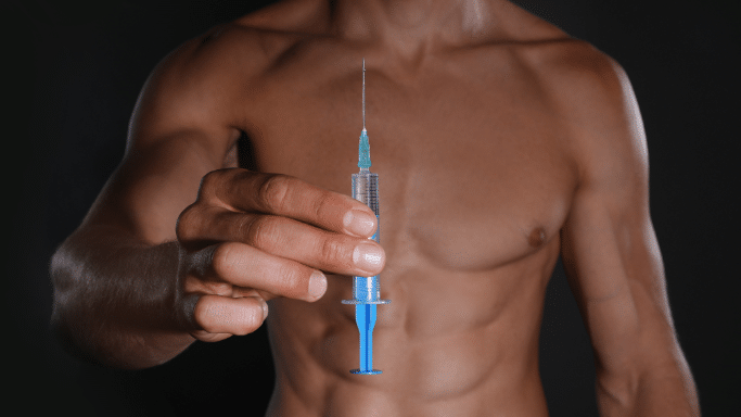 A close-up of a muscular, shirtless man's torso. He is holding a blue and clear syringe directly toward the camera, highlighting the needle and the liquid inside.