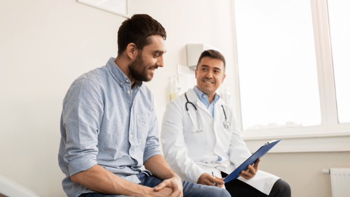 A man in a blue button-down shirt sits on an examination table in a doctor's office, smiling. A doctor in a white lab coat sits across from him, holding a clipboard and returning the smile during a consultation.