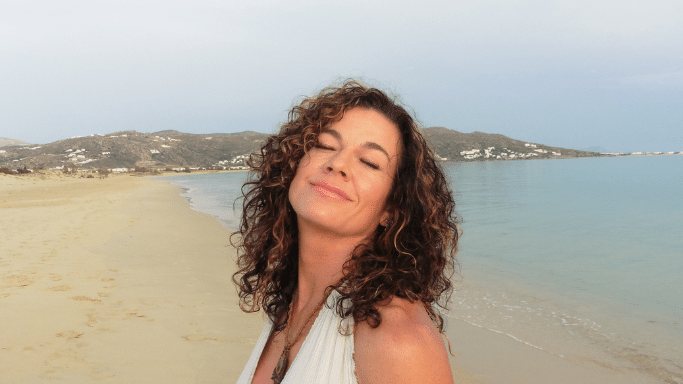 A close-up portrait of a woman with voluminous, curly brown hair with blonde highlights. She has her eyes closed and a peaceful expression, basking in the light on a sandy beach.