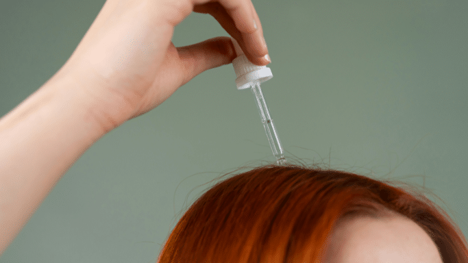A close-up shot of a person with red hair applying a clear serum to their scalp using a glass dropper.