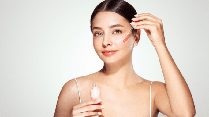 A studio portrait of a woman with her hair pulled back, applying a clear liquid from a glass dropper to her cheek. She holds a small pink bottle in her other hand.