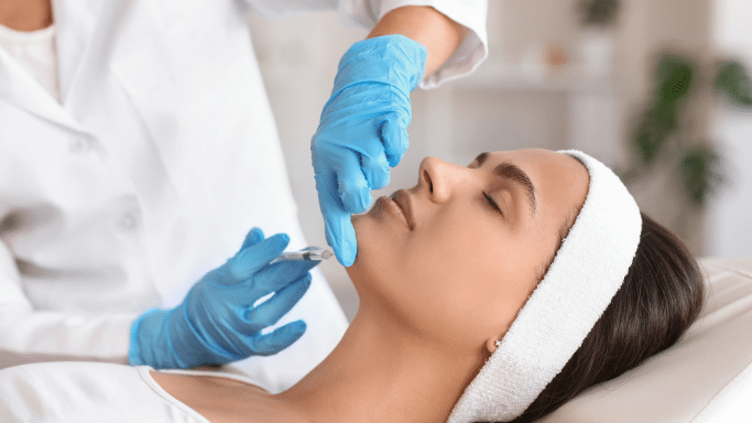 A profile view of a woman lying down receiving a cosmetic injection in her chin from a professional wearing blue nitrile gloves.
