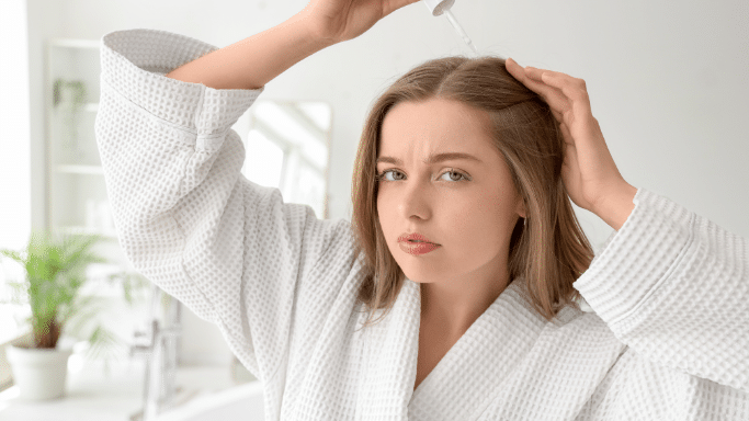 A woman in a white waffle-weave bathrobe looking into a mirror with a concerned expression while applying a treatment to her scalp with a dropper.