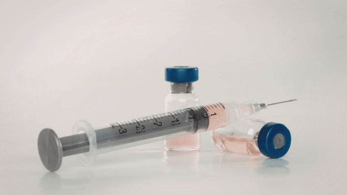 A clear medical syringe filled with a pinkish liquid lies on a white surface next to two small glass vials with blue caps.