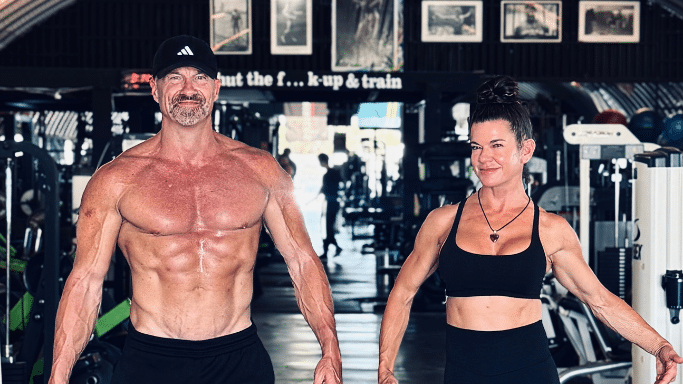 Man stands alongside a muscular woman with her hair in a bun; both are wearing black athletic gear and posing inside a gym.