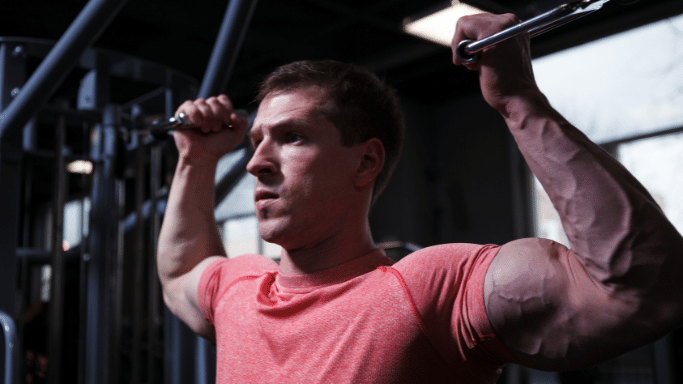A close-up, side-profile shot of a man in a red t-shirt focusing intensely while performing a pulldown exercise on a gym machine.