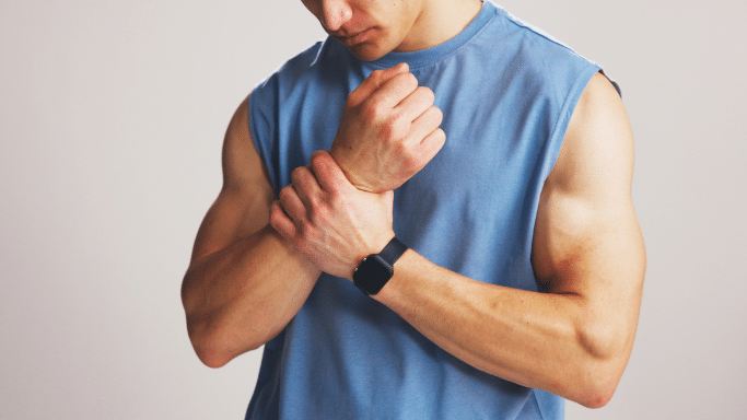 A close-up of a man in a blue sleeveless shirt holding his wrist with a pained expression, suggesting a sports-related injury or strain.