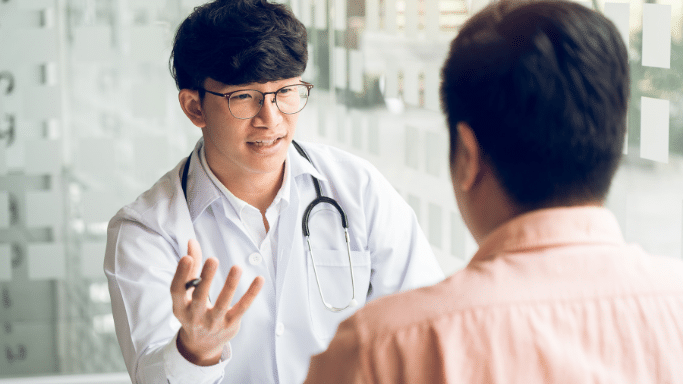 A young male doctor in a white lab coat and glasses gestures with his hand while speaking to a patient, who is seen from behind.