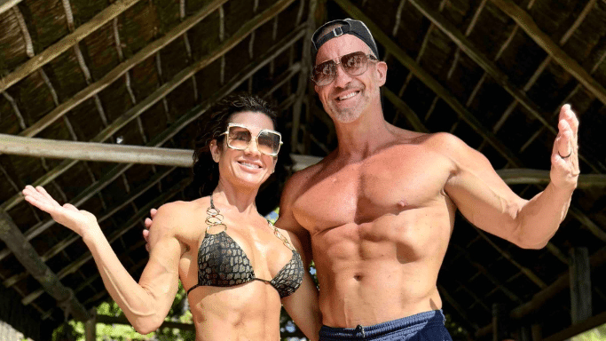 The man and woman from the gym photos pose together outdoors under a thatched roof; the woman is in a bikini and the man is shirtless, both wearing sunglasses.