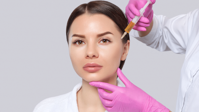 close-up of a woman receiving a facial injection. A person wearing white sleeves and bright pink nitrile gloves holds the woman's chin with one hand while using the other to aim a syringe filled with a yellow liquid toward her temple area.