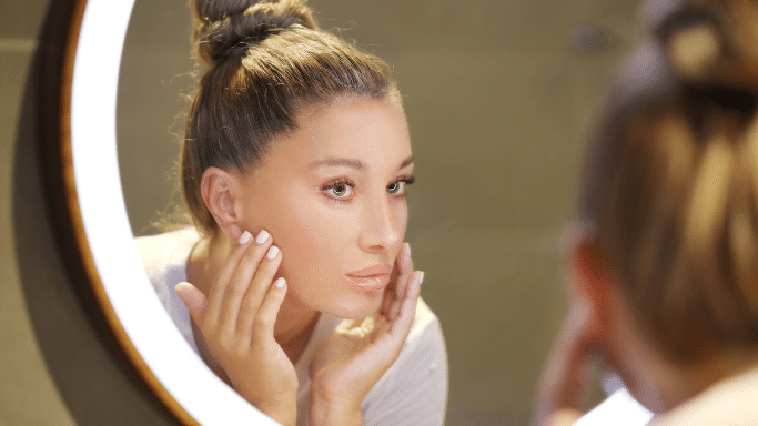 A woman with her hair in a high bun looks intently at her reflection in a circular, illuminated vanity mirror. She has her hands gently touching her cheeks, as if inspecting her skin or applying skincare.