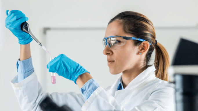 A scientist in a white lab coat, blue gloves, and protective safety glasses works in a bright laboratory. She is using a precision pipette to carefully drop a pink liquid into a glass test tube.
