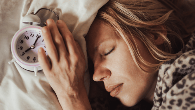 A close-up, high-angle shot of a blonde woman sleeping peacefully in bed. Her hand rests near a small, vintage-style white alarm clock on her pillow, which shows the time is just past 7:00.