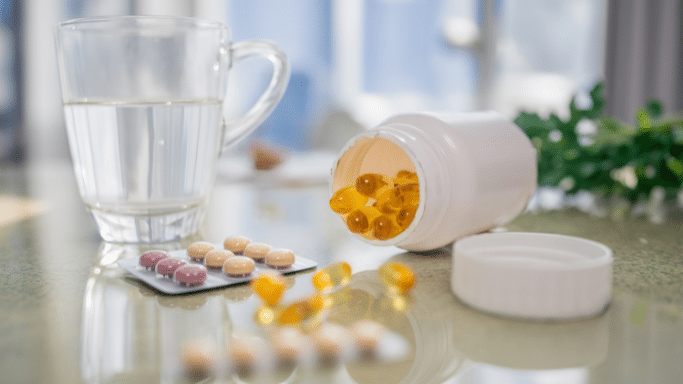 A bright, slightly blurred indoor shot of various health supplements on a table. Included are a clear glass mug of water, a blister pack of small round pills, scattered yellow gel capsules, and an open white plastic bottle spilling more capsules.