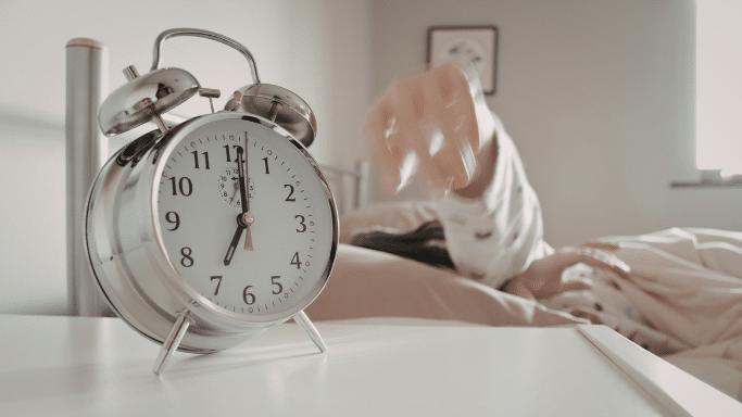 A bright bedroom scene featuring a large, silver twin-bell alarm clock in sharp focus in the foreground, showing 7:00. In the blurred background, a person in bed is reaching out a hand to turn off the alarm.