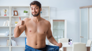 A shirtless man smiles and gives a thumbs up while pulling at the waist of oversized blue jeans to show significant weight loss.