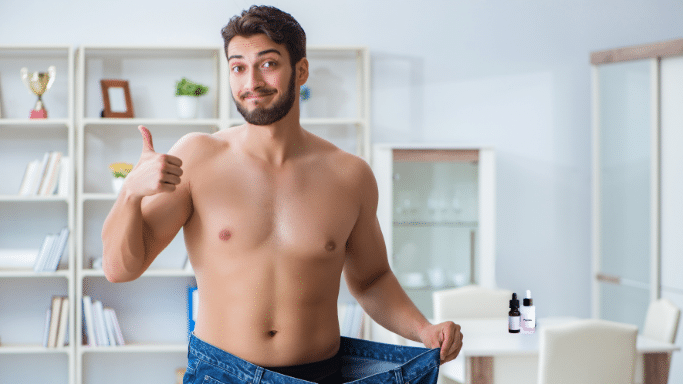 A shirtless man smiles and gives a thumbs up while pulling at the waist of oversized blue jeans to show significant weight loss.