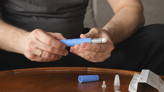 Close-up of a person’s hands preparing a blue injection pen over a wooden table with medical supplies.