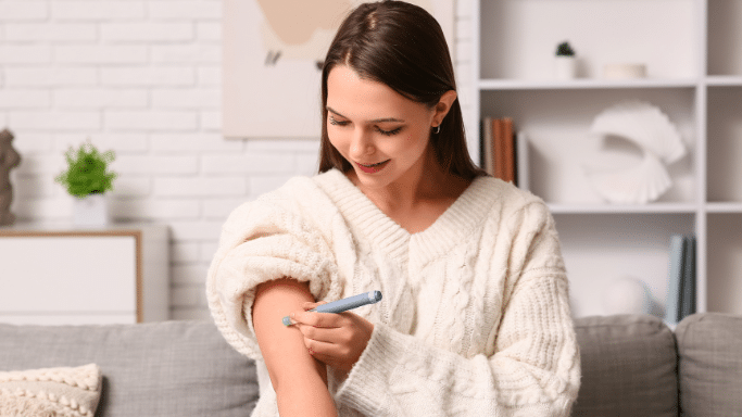 A woman in a white sweater smiling while using a medical injection pen on her upper arm in a living room.