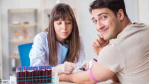 A smiling man looks at the camera while a healthcare professional in a lab coat and gloves draws his blood, with multiple vials of blood in a rack in the foreground.