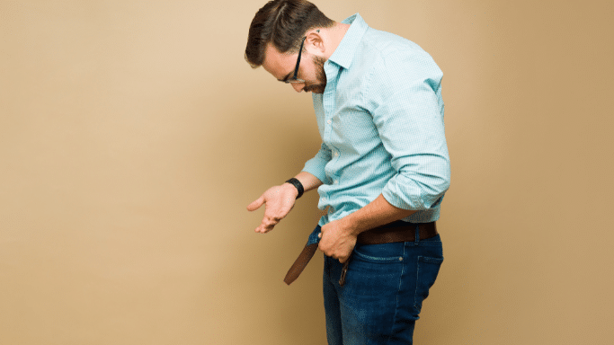 A man in a light blue shirt and jeans looks down in frustration or confusion while holding his belt and waistband away from his body.
