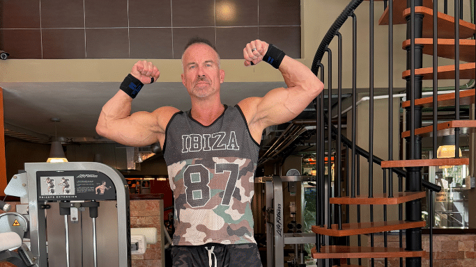 A muscular man in a gym wearing a camouflage tank top flexes both biceps while standing next to weight equipment and a spiral staircase.