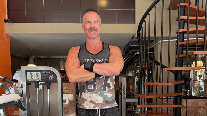 A muscular man in a gym stands with his arms crossed and a confident smile, positioned between a triceps extension machine and a wooden spiral staircase.