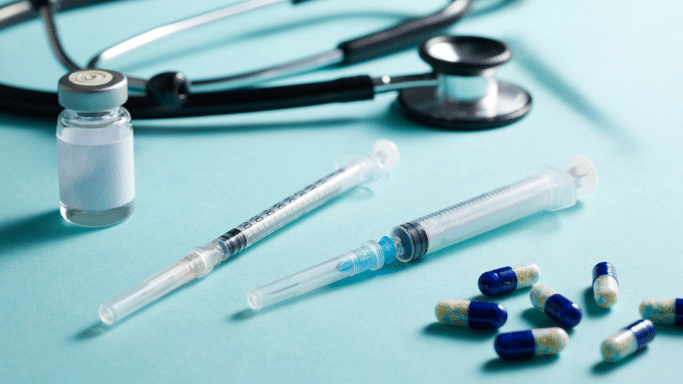 A medical-themed still life on a light blue background. It features a black stethoscope, a small glass vial with a white label, two plastic syringes with needles capped, and a scattered handful of blue and white speckled capsules.