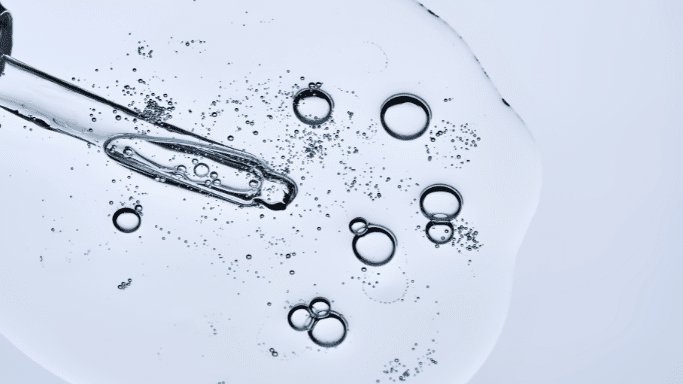 A high-angle, close-up shot of a clear, viscous liquid (like a facial serum) spread across a white surface. A glass dropper is submerged in the liquid, surrounded by various sizes of air bubbles.