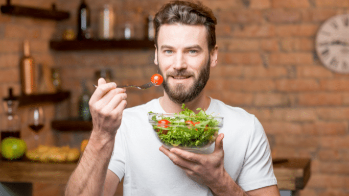 A smiling man with a beard stands in a kitchen, holding a glass bowl of green salad with cherry tomatoes. He is holding a fork with a single cherry tomato up toward his mouth as if about to eat.